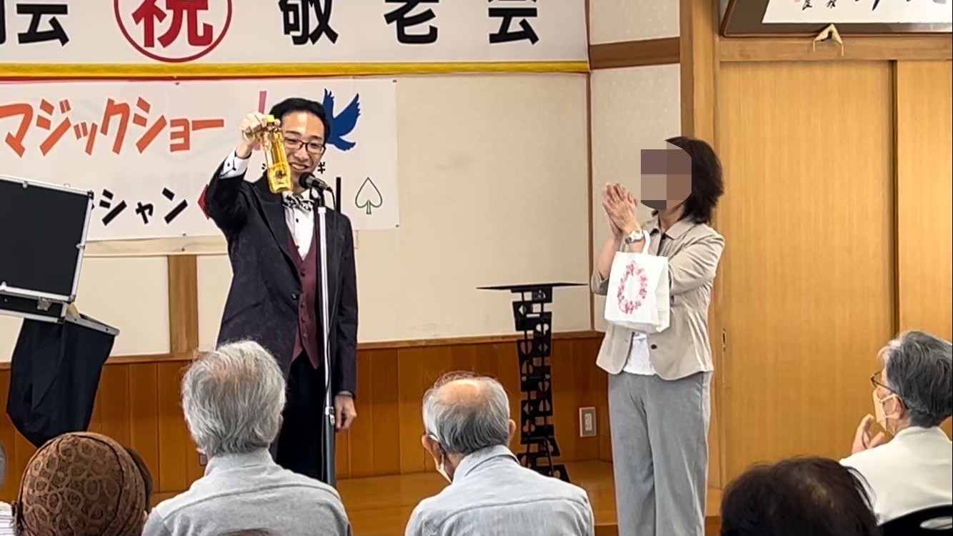 「祝 敬老会 マジックショー」の横断幕の前で、客席に向かってマジックを見せているマジシャンSHIGI